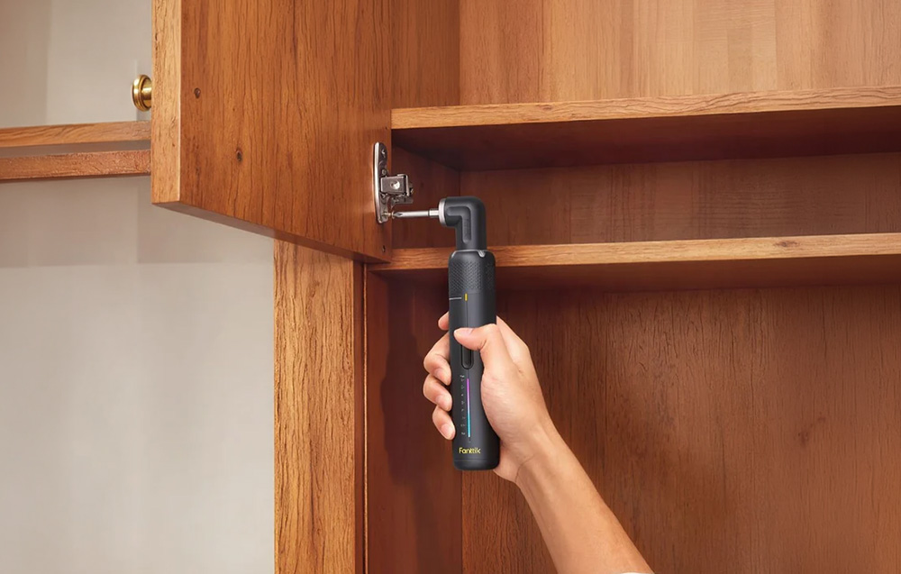 A cordless screwdriver with an angle attachment screws a hinge into the interior of a wooden cabinet