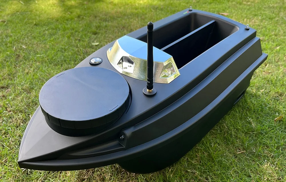 A black bait boat with two LED compartments, an antenna, and a round hopper, resting on green grass