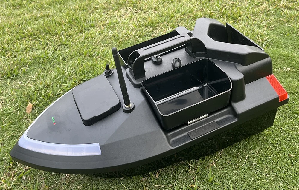 Black RC bait boat with empty container and red accent on the back, lying on green grass