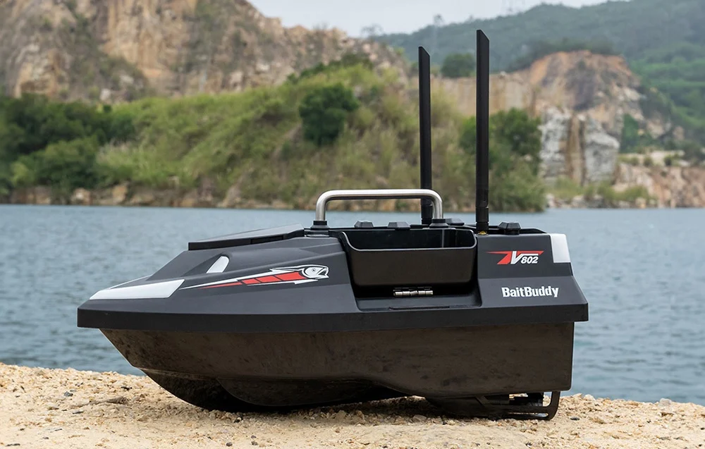 RC BaitBuddy V602 bait boat with two antennas standing on the lake shore with mountains in the background.