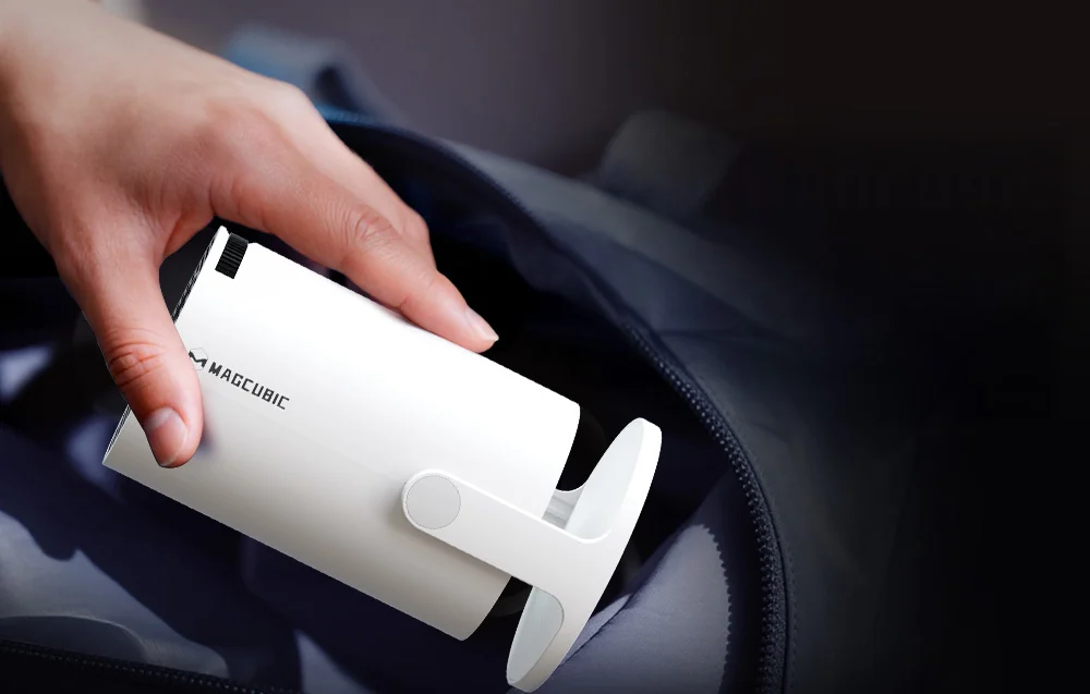 A hand places a compact, white Magcubic projector into an open backpack or travel bag