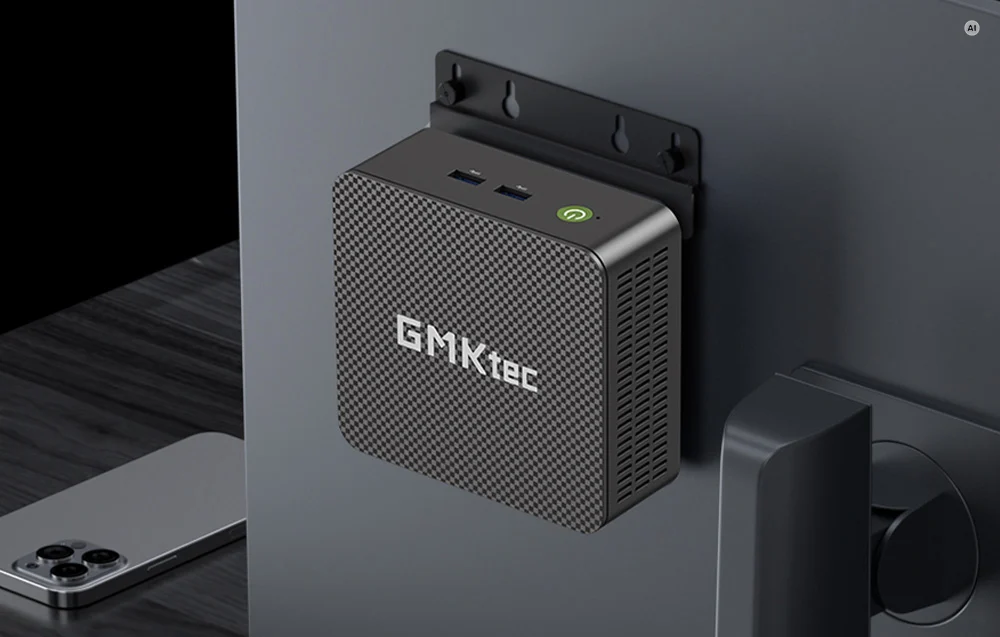 A GMKtec mini PC mounted on the back of a monitor using a mounting bracket, with a smartphone visible next to it.