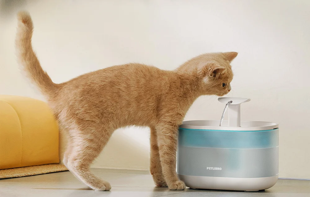 A ginger cat drinking water from an automatic water fountain standing next to a wall.
