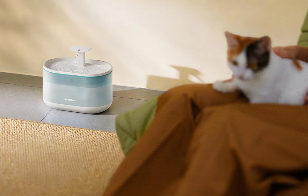 A cat lying next to an automatic water fountain placed on the floor.