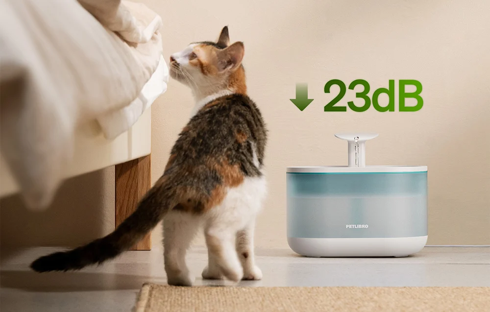 A tricolor cat stands next to an automatic water dispenser placed on the floor next to the bed.