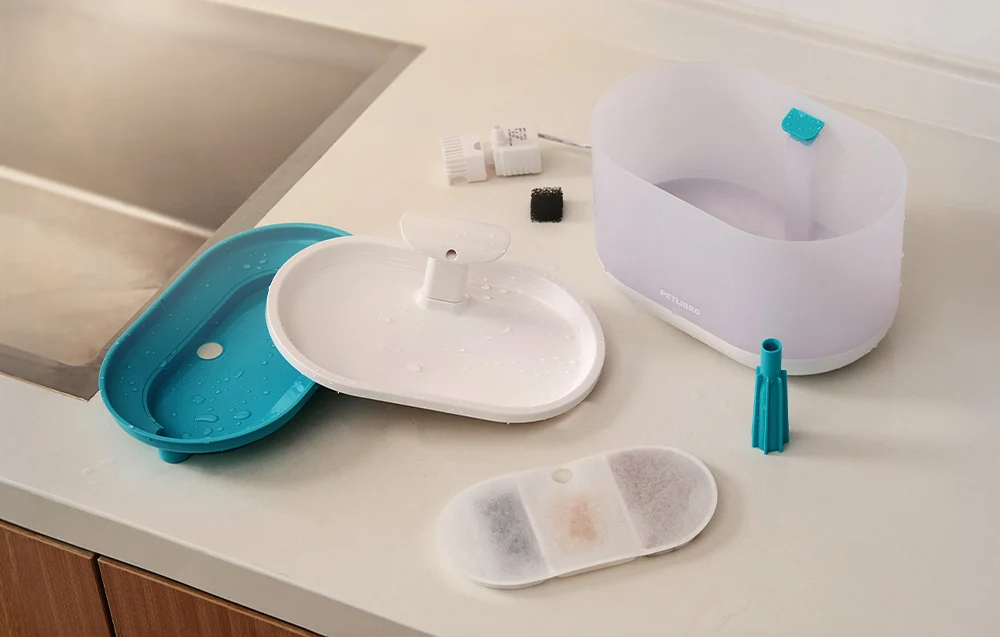 The components of a cat water dispenser laid out on a kitchen counter, including a water reservoir, a plastic lid, a filter, and cleaning accessories.