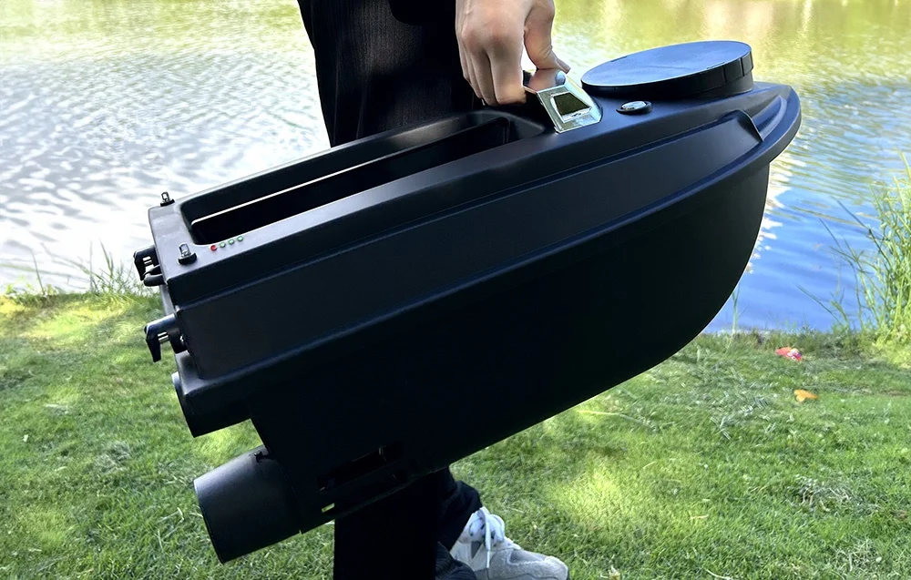 A person holding a black bait boat above the water, touching the battery level indicator