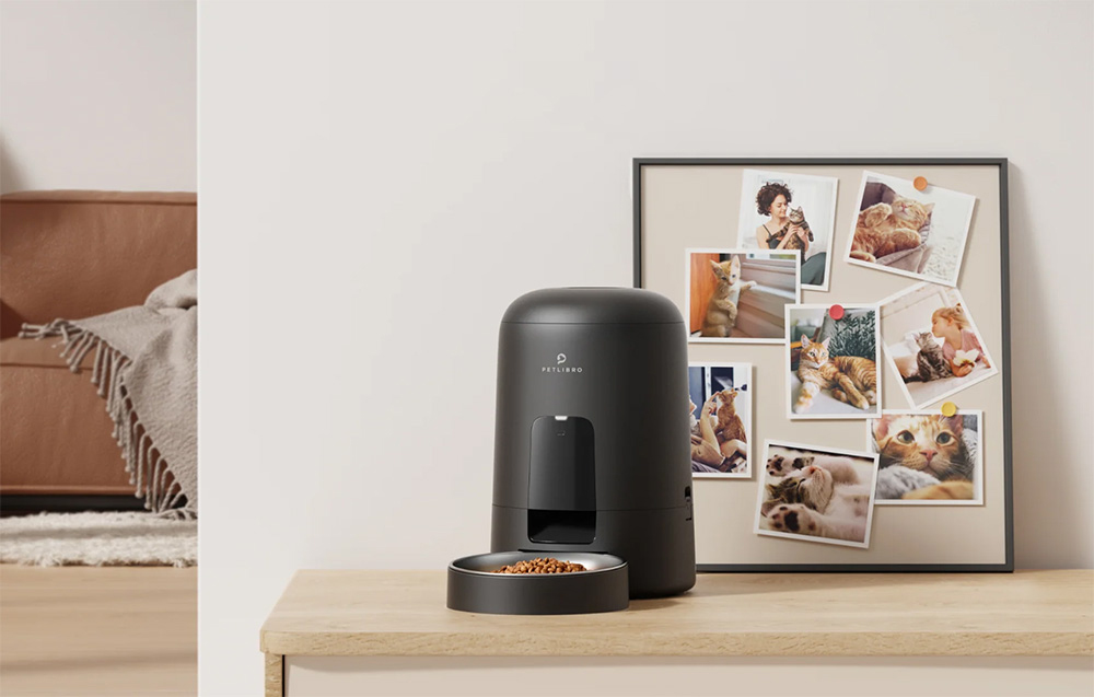A black automatic feeder with a bowl full of dry food stands on a wooden cabinet next to a photo frame with pictures of a cat