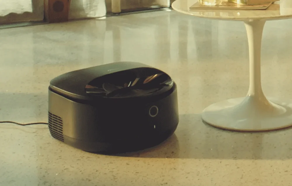 A black automatic feeder stands on a light-colored floor next to a white table