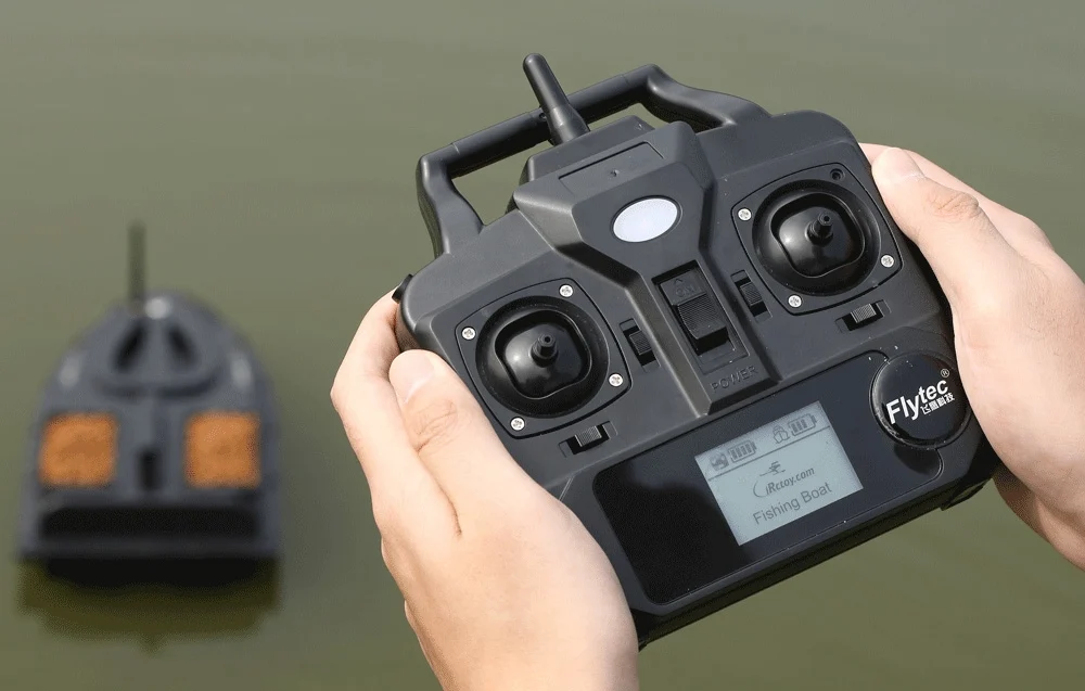 Close-up of a Flytec remote control with a "Fishing Boat" display, with a blurred bait boat on the water in the background