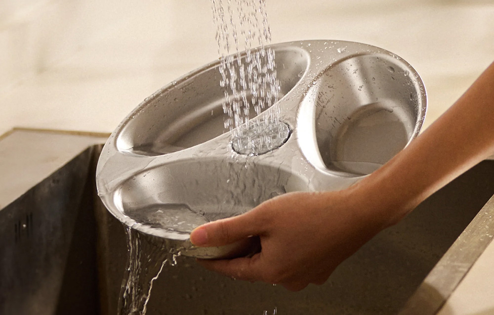 A hand holds a three-compartment bowl insert under running water at the sink
