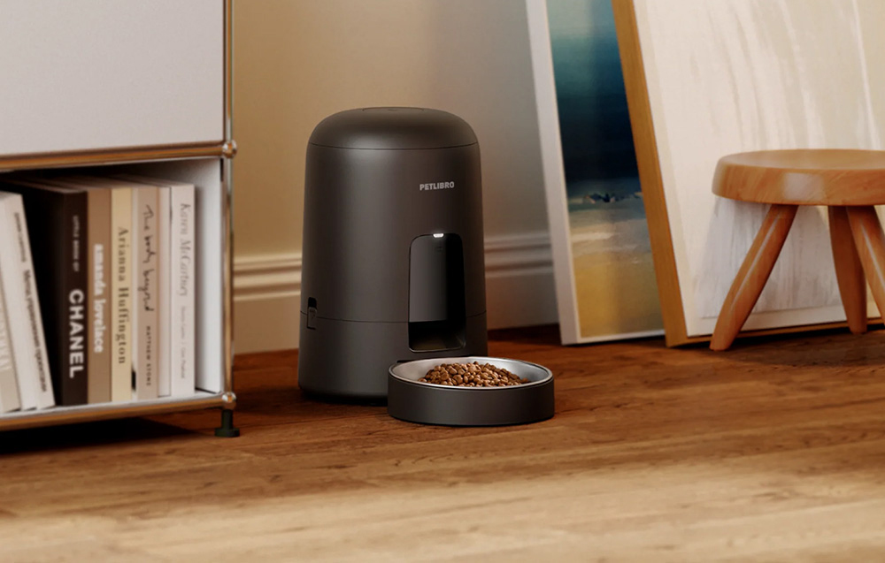 A black automatic feeder with a bowl full of dry food stands on a wooden floor next to a bookshelf and a stool