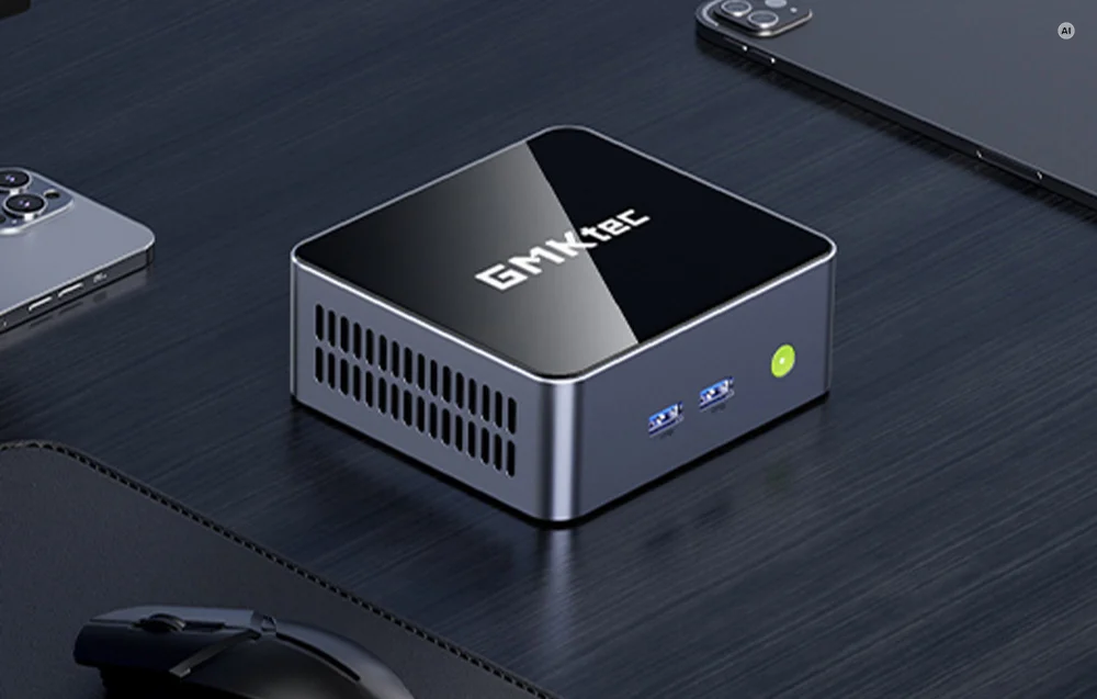 A gray GMKtec mini PC resting on a dark desk next to a smartphone, tablet, and mouse.