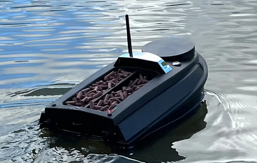 A black bait boat with an open compartment filled with dark bait, floating on the water