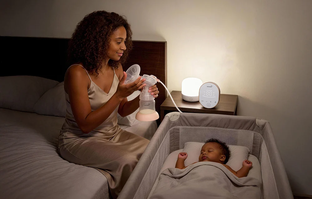 A woman is sitting by the bed using a breast pump while her baby sleeps in the crib next to her