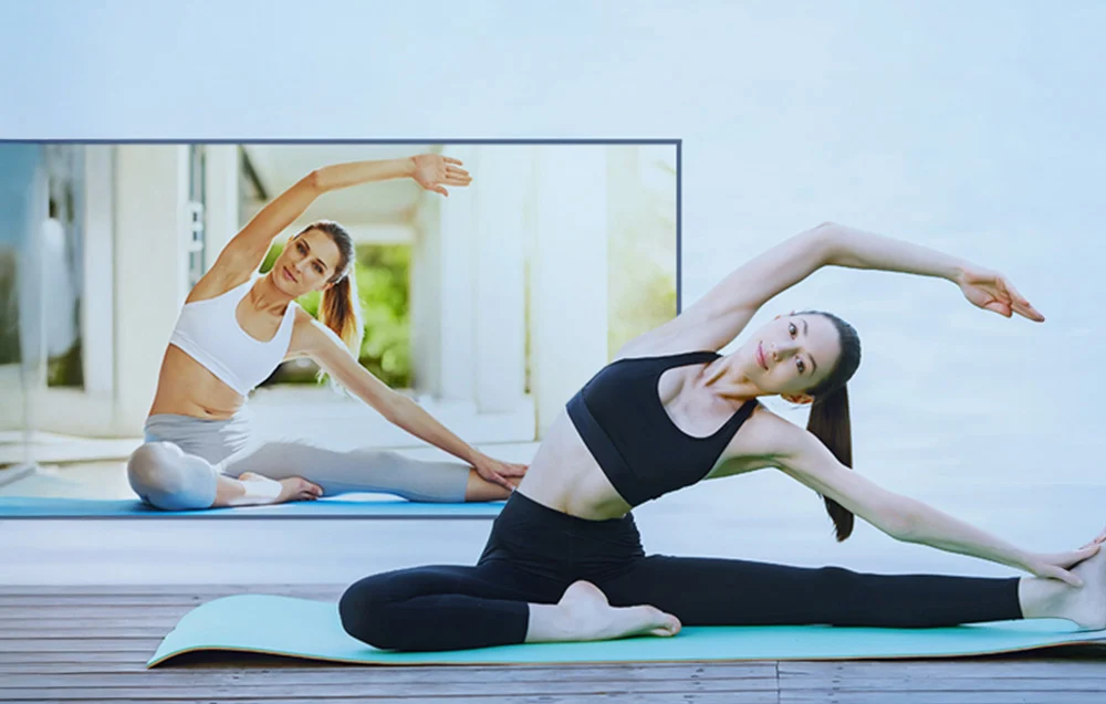 A woman practices yoga on a mat, mimicking the movements of an instructor visible in a large image projected onto the wall