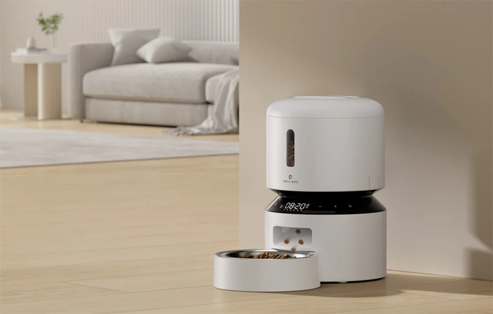 A white automatic cat feeder stands on the living room floor and dispenses food into a bowl
