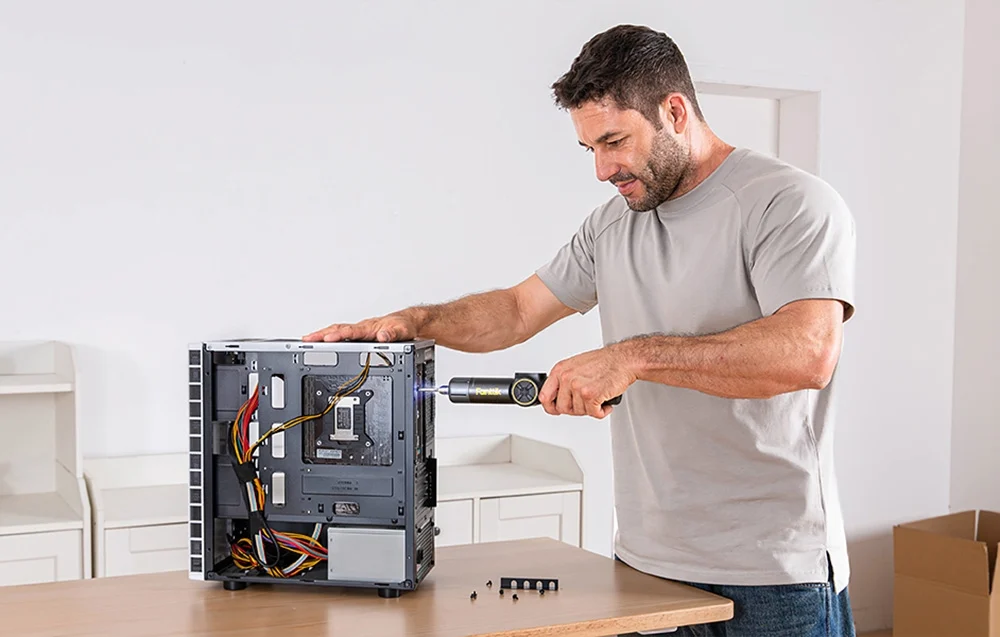 A man in a gray T-shirt and jeans repairs a computer with an electric screwdriver.