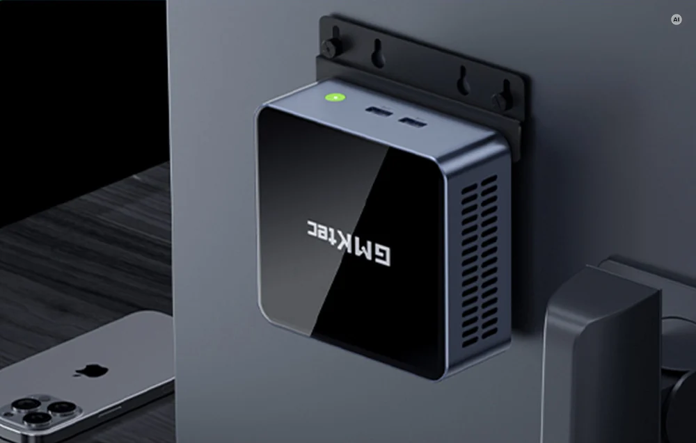 A GMKtec mini PC mounted on a VESA mount on the back of a monitor next to a smartphone.