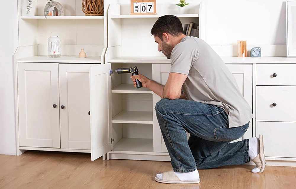 A man in a gray shirt screws a cabinet door with an electric screwdriver.