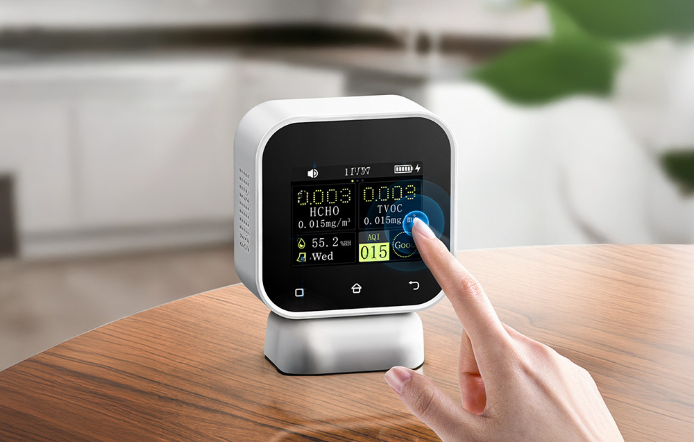 A hand touching the screen of an air quality monitor standing on a wooden table.