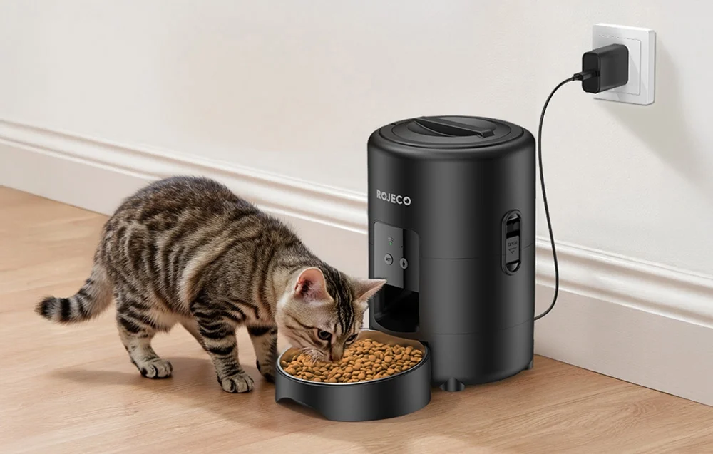 Cat eating from black food dispenser plugged in against wall